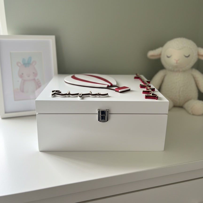 White baby keepsake box with a red and white balloon design on a white surface with a lamb plush toy and framed picture in the background.