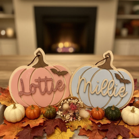 Decorative pumpkin-shaped signs with names 'Lottie' and 'Millie' on a table with fall leaves and pumpkins, blurred fireplace in the background.