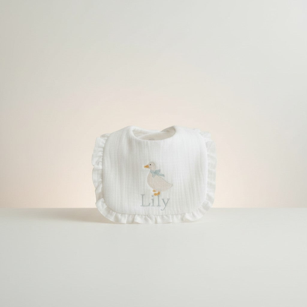 The Wee Rhubarb Gift Co Ltd white muslin baby bib with embroidered duck, blue bow, ruffles and name 'Lily'.