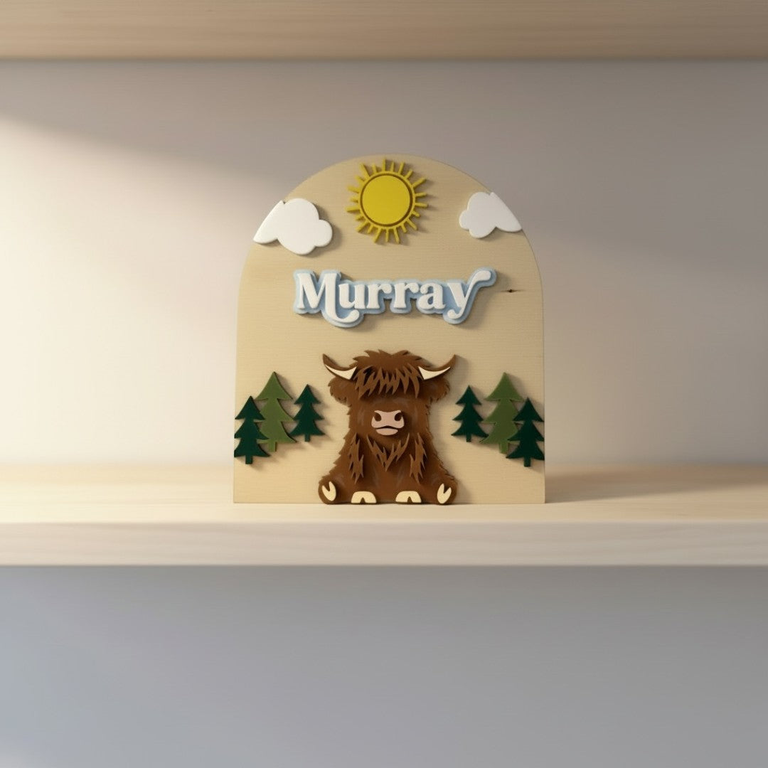 The Wee Rhubarb Gift Co Ltd Highland Cow Wooden Name Plaque with 'Murray' in blue layered font, sun, clouds, and trees.