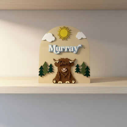 The Wee Rhubarb Gift Co Ltd Highland Cow Wooden Name Plaque with 'Murray' in blue layered font, sun, clouds, and trees.