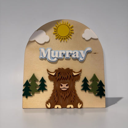 The Wee Rhubarb Gift Co Ltd Highland Cow Wooden Name Plaque with 'Murray' in blue layered font, sun, clouds, and trees.