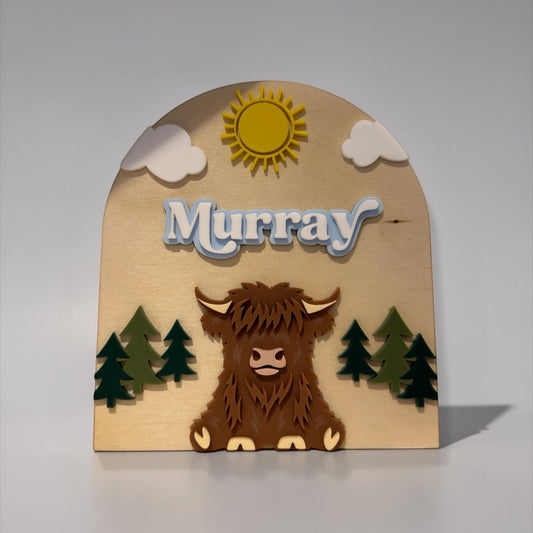 The Wee Rhubarb Gift Co Ltd Highland Cow Wooden Name Plaque with 'Murray' in blue layered font, sun, clouds, and trees.
